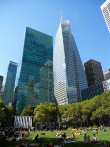 People lounge on green grass in Bryant Park, Manhattan, surrounded by glass skyscrapers beneath a clear blue sky.