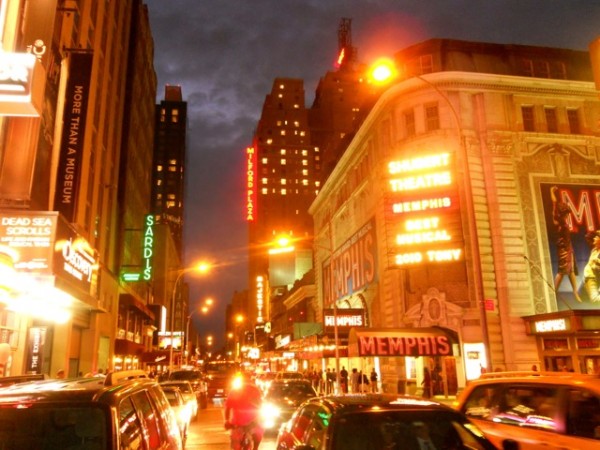 A bustling New York City street at night features Broadway theater marquees for “Memphis,” vibrant neon signs, and dense crowds.