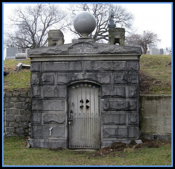 A stone mausoleum with “Jonathan Reed” and “Mary E. Reed” engraved stands in a wintry cemetery bordered by leafless trees.