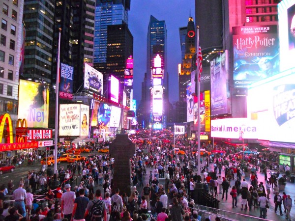 A large crowd gathers in Times Square, New York City at dusk. Bright billboards and yellow taxis reflect city nightlife.