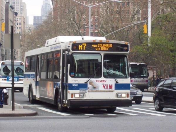 A blue and white M7 bus travels through a busy Manhattan intersection near tall buildings, with trees showing sparse autumn leaves.