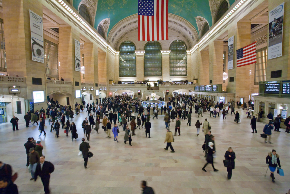A busy crowd moves through Grand Central Terminal’s main hall in New York City, beneath arched windows and American flags.