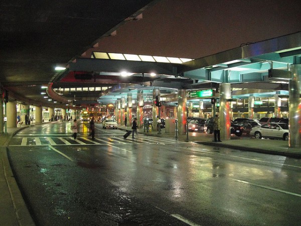 Several people walk beneath a canopy of lights and metal beams outside a New York airport terminal on a wet night.