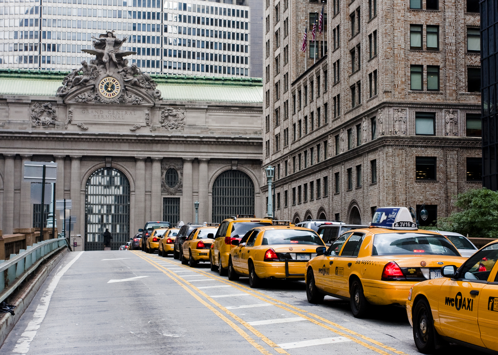 Several yellow taxis are parked in a line outside Grand Central Terminal in New York City, surrounded by tall office buildings.