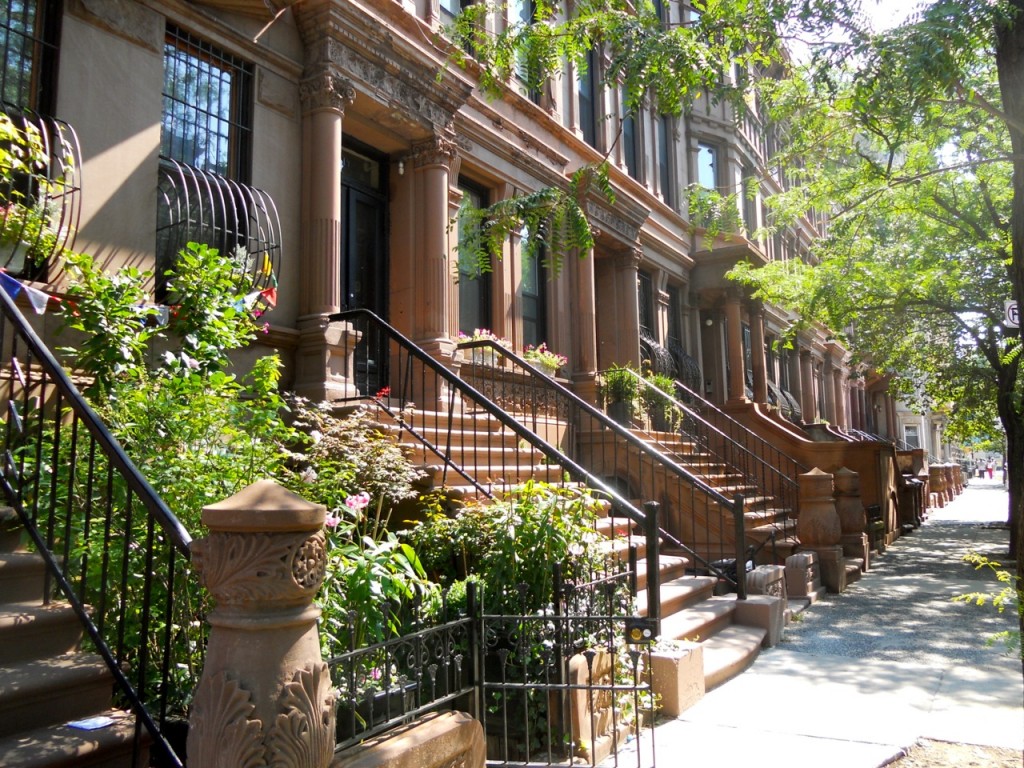 Classic brownstone buildings with stoops and wrought-iron railings line a sunny, tree-lined Harlem sidewalk, celebrating NYC Black heritage.