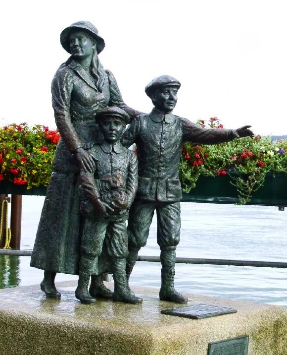 A bronze statue of Annie Moore and two boys stands near Ellis Island, with one boy pointing forward and water behind them.