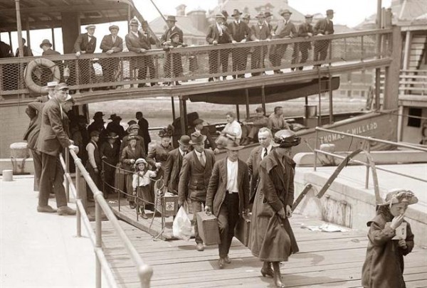 Several people in early 20th-century clothing walk along a pier toward a docked boat, observed by others from an upper deck.