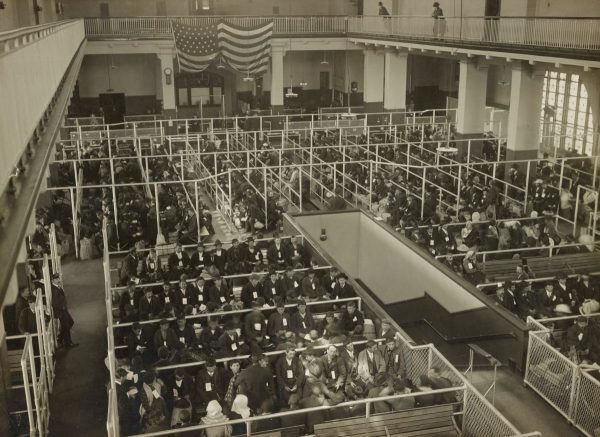A large group of immigrants sits in sectioned waiting areas beneath an American flag inside Ellis Islands main hall.