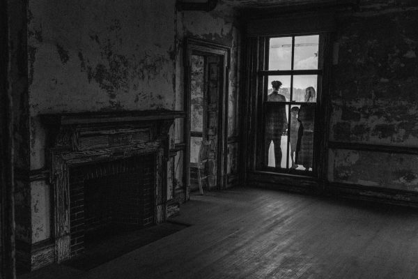 A dim, empty room with peeling walls and fireplace; outside the window, three shadowy figures stand silhouetted in eerie light.