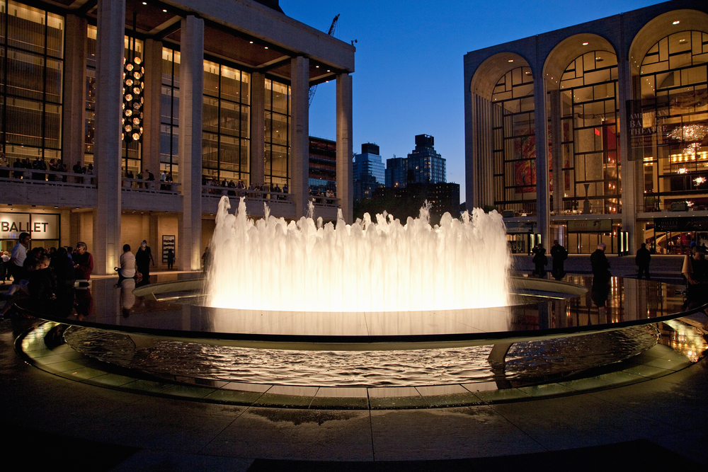People gather around the illuminated fountain at Lincoln Center, New York, as modern glass buildings glow against the evening sky.