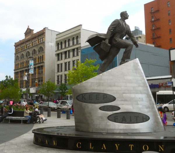 A bronze statue of a suited man holding his coat stands on a “KEEP THE FAITH” pedestal in Harlem, surrounded by people.