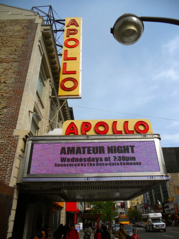 The Apollo Theater’s bright yellow “APOLLO” sign and marquee advertises “Amateur Night Wednesdays” above a bustling Harlem street.