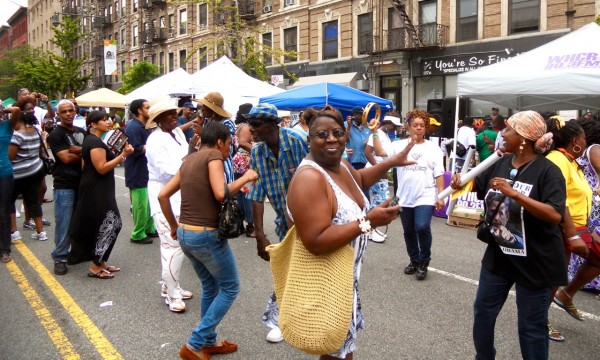 A joyful crowd dances at a Harlem street festival, surrounded by tents, music performers, and city buildings on a sunny day.