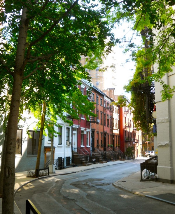 A peaceful Greenwich Village street curves ahead, bordered by colorful brownstones, leafy trees, and black iron railings under warm sunlight.