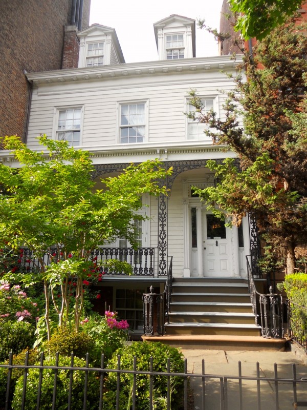 A white two-story house in Brooklyn Heights features intricate iron railings, a flower-filled garden, and two rooftop dormer windows.