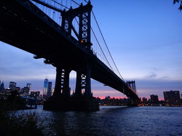 The Brooklyn Bridge stretches over the East River at dusk, with Manhattan skyscrapers glowing against a vivid purple and pink sky.