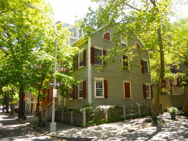 A gray house with red shutters stands on Milligan Street in Brooklyn Heights, surrounded by trees, greenery, and a low fence.