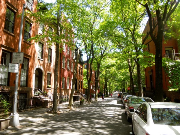Sunlit Brooklyn Heights street lined with historic brownstones, leafy trees, parked cars; pedestrians stroll beneath dappled shadows on the sidewalk.