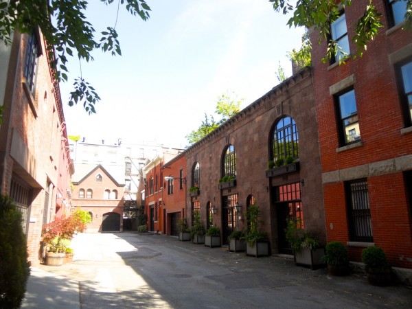 A peaceful alley in Brooklyn Heights features red brick buildings, arched windows, potted plants, and leafy trees on a sunny afternoon.