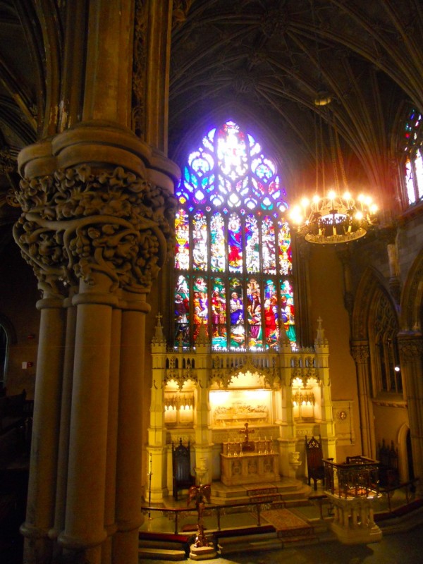 A grand church sanctuary in Brooklyn Heights displays intricate stone columns, a luminous chandelier, and colorful stained glass above the altar.