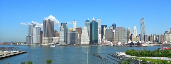 The Manhattan skyline rises behind the East River as a ferry passes, with Brooklyn Heights greenery and clear blue sky visible.