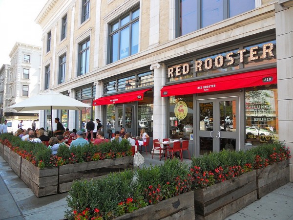 People enjoy lunch under red awnings at Red Rooster in Harlem, surrounded by flower planters and a large beige building.