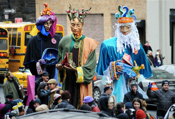 Three performers on stilts wear vibrant, whimsical giant puppet costumes, entertaining a bustling crowd at New York Citys Three Kings Day parade.