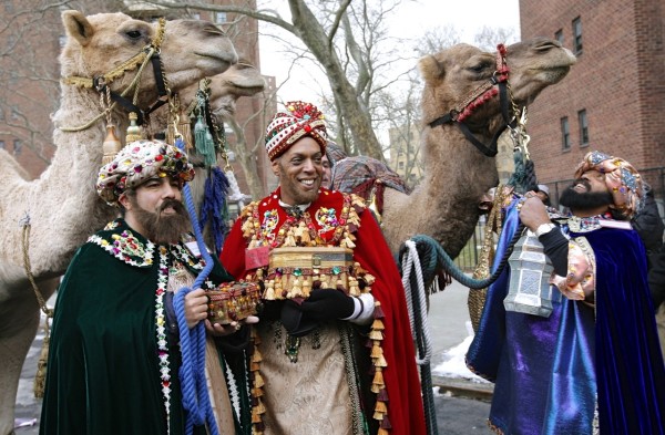 Three people dressed as the Three Wise Men stand near camels and brick buildings in New York City during Three Kings Day festivities.
