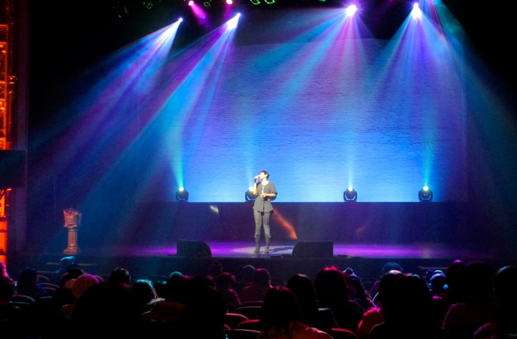 A singer performs under colorful spotlights on the Apollo Theater stage in Harlem, while an attentive audience watches from dimly lit seats.