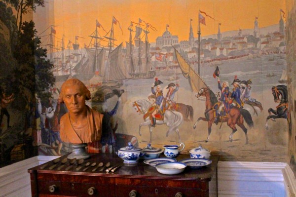 A bust of George Washington sits on a dresser beside blue and white dishes, with a mural of New York’s Revolutionary era.