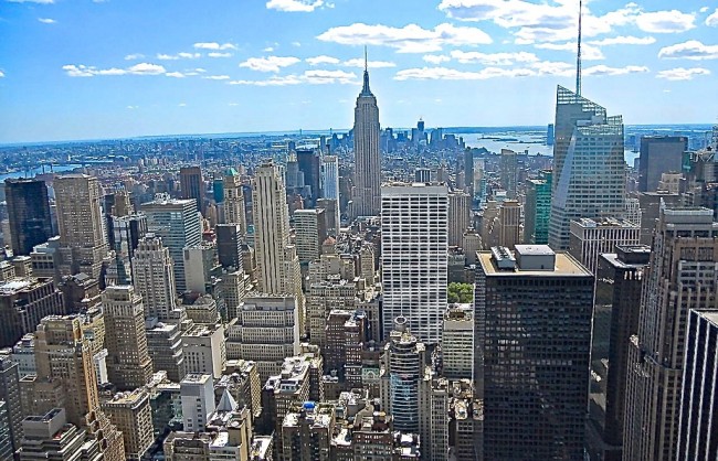 Tall skyscrapers surround the Empire State Building in Midtown Manhattan under a bright blue sky with scattered white clouds.