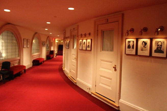 A corridor of a theatre, curving behind the auditorium. A red carpet seperates the white walls, with arched windows on the left and white doors entering the auditorium on the left, interspersed with black and white photos of famous performers.