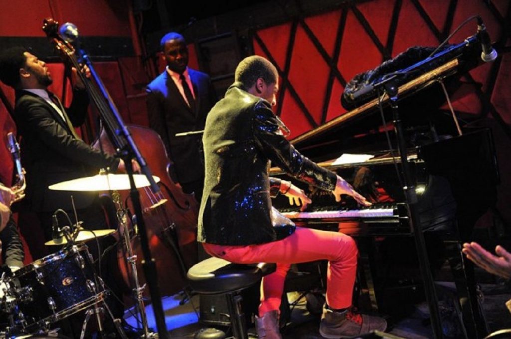 A jazz band plays energetically onstage in Harlem, with a sequined pianist, double bassist, and drummer captivating an enthusiastic audience.