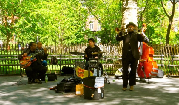 A jazz band plays in a sunny West Village park, with instruments and open cases for donations while people walk by.