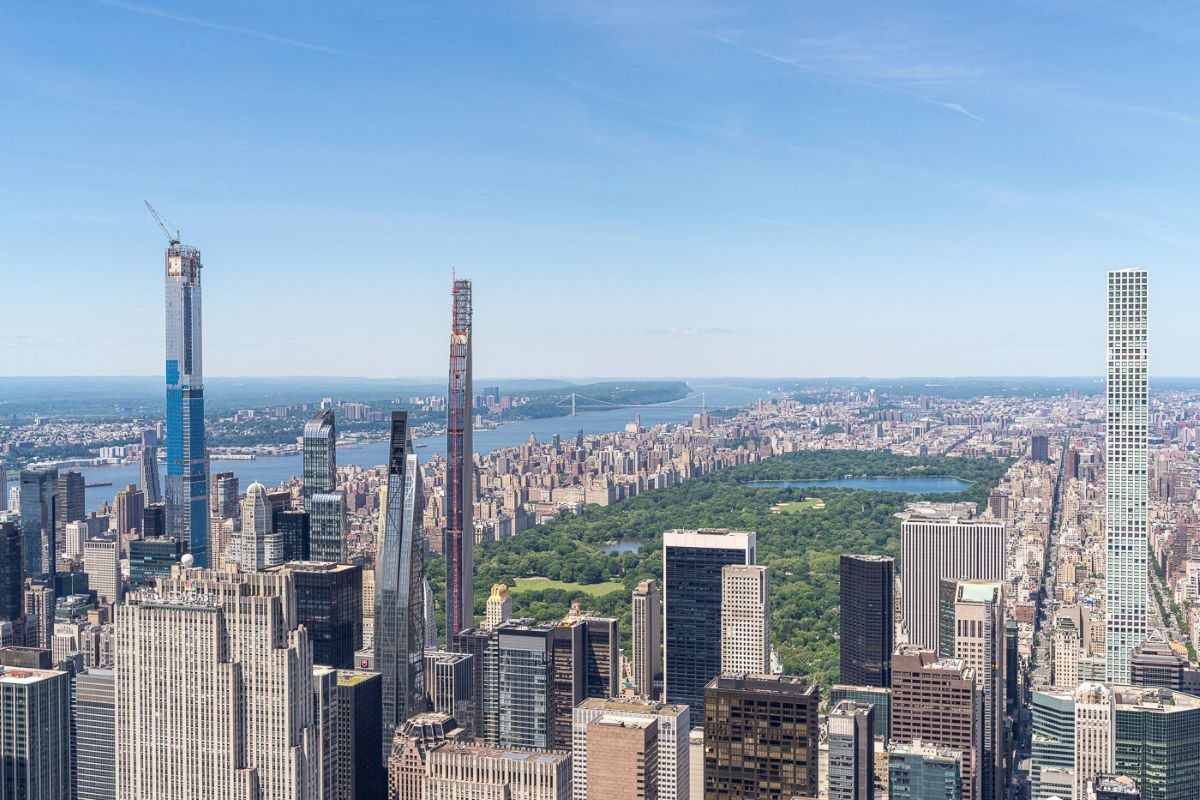 Skyscrapers of Midtown Manhattan surround Central Park, with the Hudson River and cityscape visible under a clear blue sky.