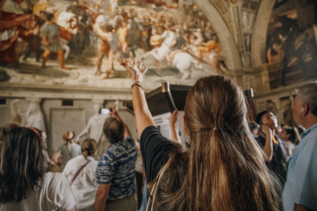 A woman photographs Diego Riveras vibrant mural in a packed museum room, while others admire and document the famous artwork.
