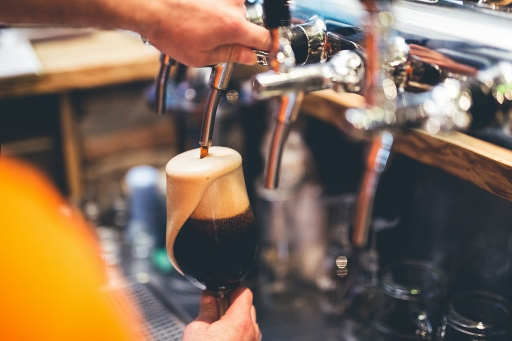 A person pours dark beer from a shiny tap, forming a thick head atop the glass on a wooden pub counter.
