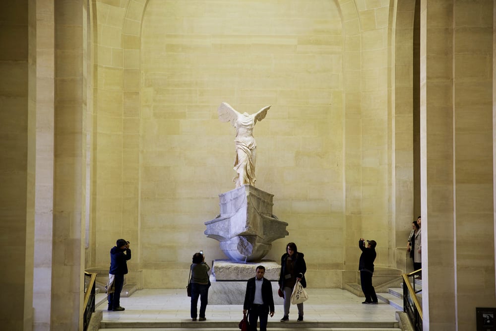 People gather around the Winged Victory of Samothrace statue at the Louvre, taking photos under tall beige walls and sunlight.