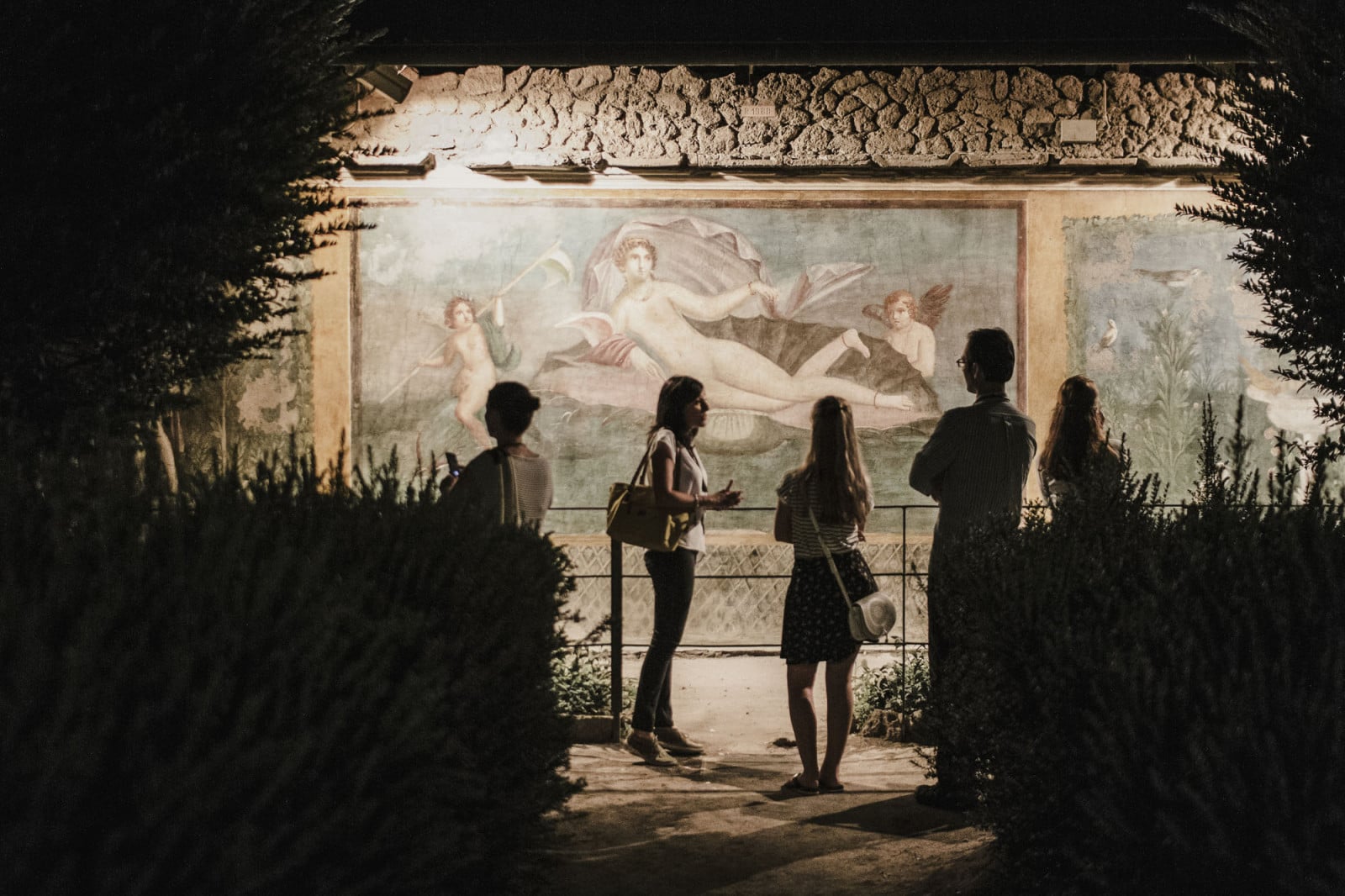 Five people stand at night in front of a brightly lit mural showing mythological gods, with trees and shadows around them, listening to their Pompeii travel guide