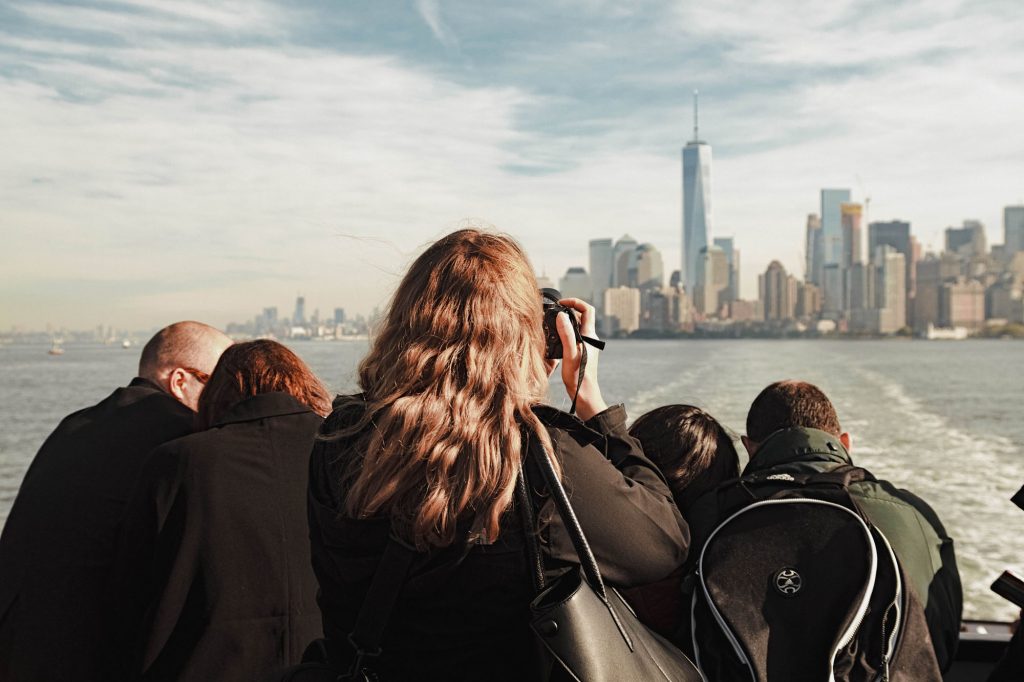 A group on a boat tours New York Harbor; one person photographs the skyline with One World Trade Center in view.