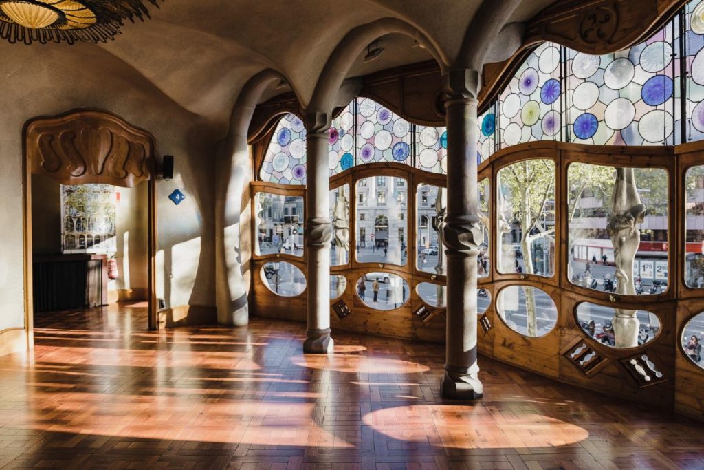 Inside Casa Batllo showing beautiful stained glass windows, curved ceilings, and tree-like pillars. Sunlight pours in through the windows, offering a glimpse of Barcelona's prominent street, Passeig de Gràcia.