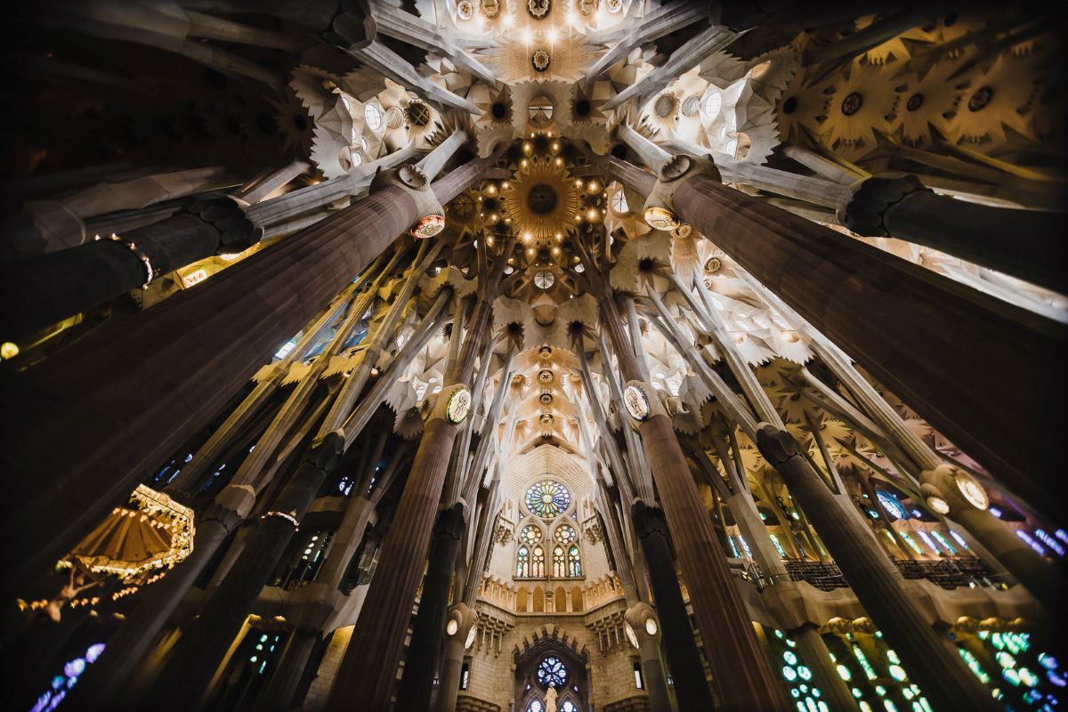 The ornate ceiling of Sagrada Familia in Barcelona features illuminated stained glass, geometric patterns, and tall columns designed by Antoni Gaudí. The most famous Gaudi building barcelona.