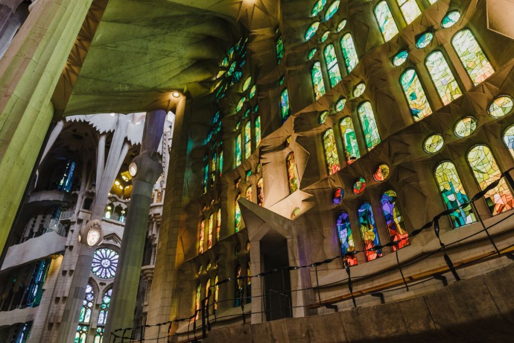 Sunlight streams through tall stained glass windows in Chartres Cathedral, coloring the stone walls and highlighting intricate Gothic architectural details.
