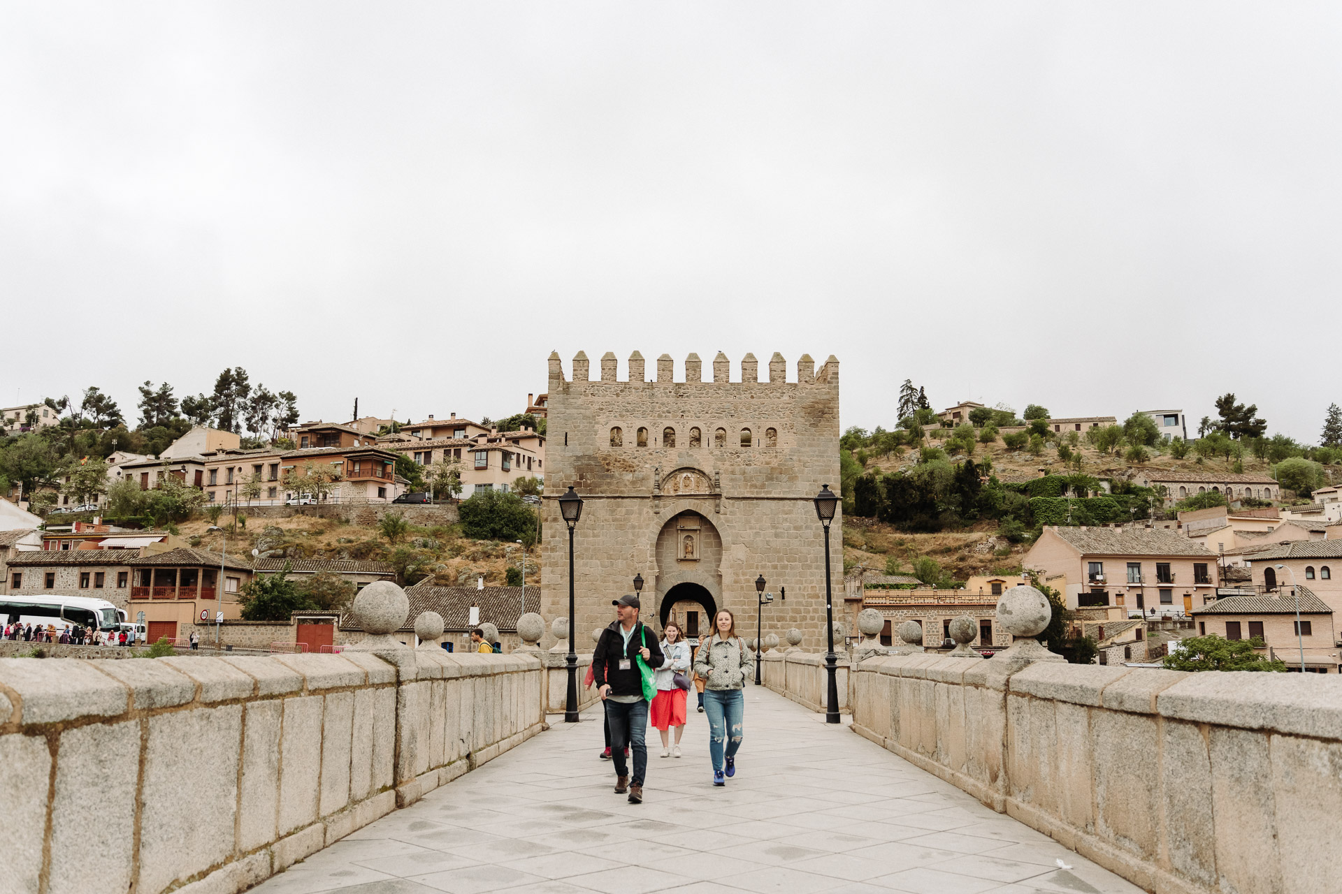 Three people cross a stone bridge toward the medieval Puerta de Alcántara gate in Toledo, Spain, surrounded by historic architecture and trees.
