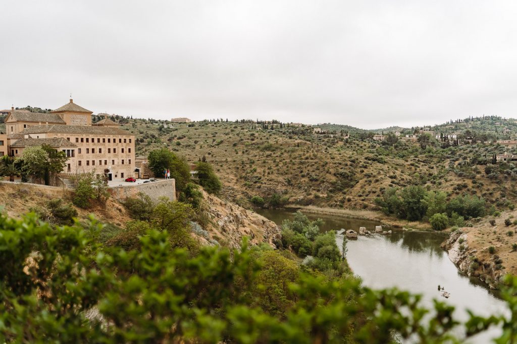 A view from the Toledo Day Trip and Winery Tour. A grand historic building stands atop a hill, overlooking a winding river and green, sparsely wooded hills under cloudy skies.