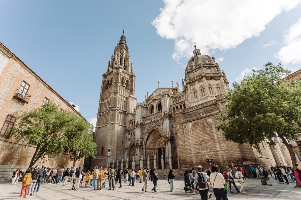 Walks_Madrid_Toledo Day Trip WB-0023 A large crowd stands outside Toledo Cathedral, admiring its ornate stone façade and tall towers under a bright blue sky.