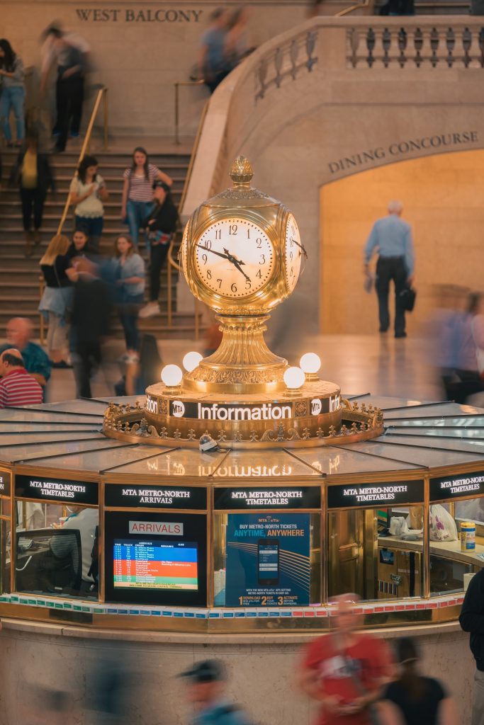 A gold clock sits on the information booth in Grand Central Terminal’s main hall as crowds pass and signs point to balconies.