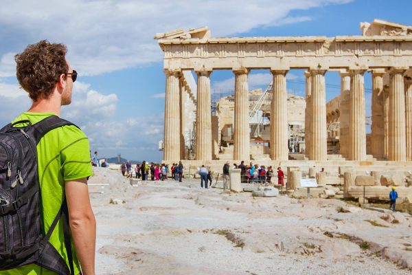 A man with a backpack stands near the Parthenon in Athens, watching tourists gather by the ancient ruins under a partly cloudy sky.