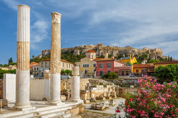 Ancient marble columns and ruins in Athens, Greece. The Acropolis rises behind colorful buildings and blooming pink flowers under a partly cloudy sky.
