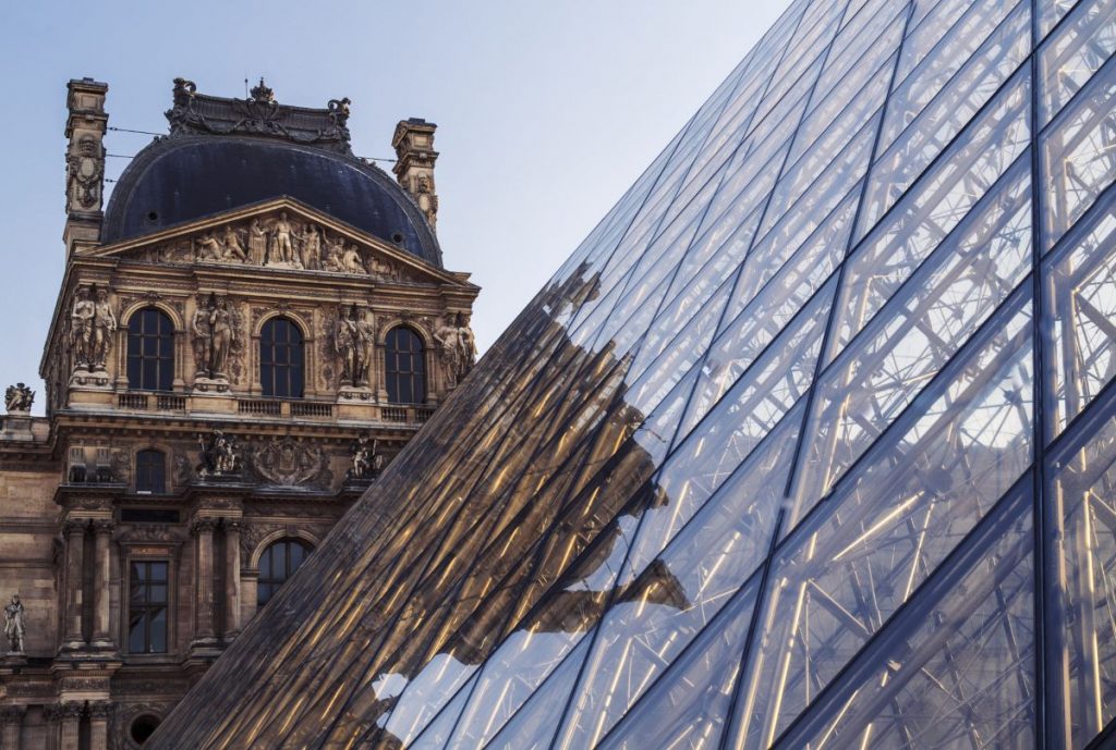 The glass pyramid at the Louvre Museum in Paris reflects ornate sculptures and historic architecture under a bright, clear sky. The best time to visit the Louvre is also the most photogenic